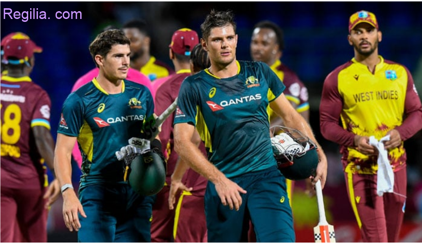 Australian cricketers walk off the field after a T20I match against West Indies, holding their helmets and gear, with West Indies players in the background.