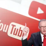 A serious-looking man in a suit and red tie gestures with his finger raised, standing in front of a large YouTube logo backdrop.