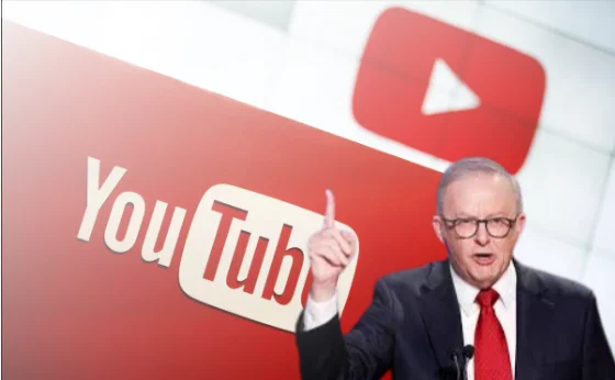 A serious-looking man in a suit and red tie gestures with his finger raised, standing in front of a large YouTube logo backdrop.