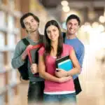 Three university students standing in a library holding books and smiling at the camera.