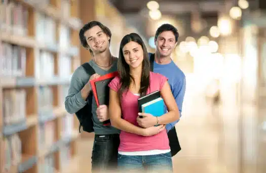Three university students standing in a library holding books and smiling at the camera.